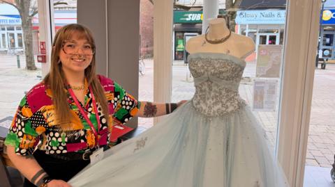 A woman smiles as she holds a princess dress, which is baby blue with glittery embellishments, on a mannequin in a shop window, with a high street outside. The woman wears large glasses and a bold-patterned multi-coloured shirt. She has prominent facial piercings. 