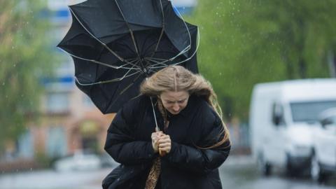 A woman in a large black puffer coat is holding a black umbrella which is turned inside out. The woman is looking down as she is being rained on. In the background are blurred images of vehicles and a street.