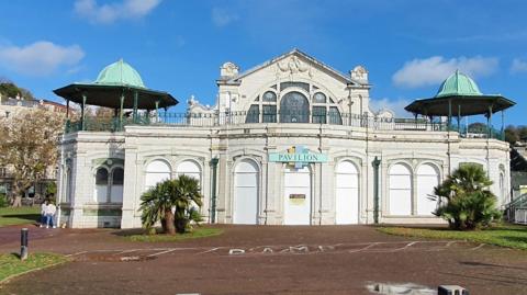 Torquay Pavilion exterior. The large building is light-grey in colour with a blue pavilion sign. Grass areas are in front of the building. 