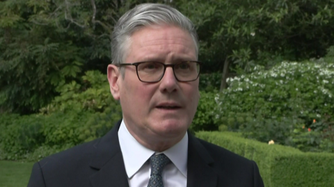 A headshot of Prime Minister Sir Keir Starmer, who is wearing a black jacket,a white shirt and a green tie. He is standing in a garden, about to go into talks on Iran. 