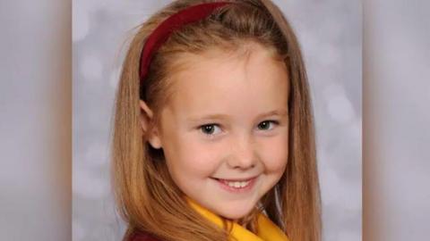 Elsie Dot Stancombe, wearing a red hairband in her long fair hair and her school uniform, smiling at the camera in a school portrait
