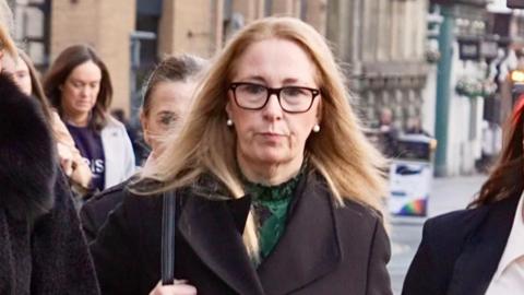 Kelly, wearing glasses and a black coat, walking up a street surrounded by other women in smart clothing