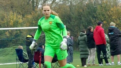 Amelia Aplin has long blonde hair and is wearing a bright green goalkeeper kit with white gloves.