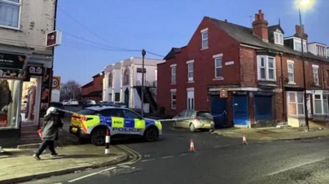 A road is closed off with cones in the road and police tape across the top of the street. There is a marked police car parked across the top of the street 