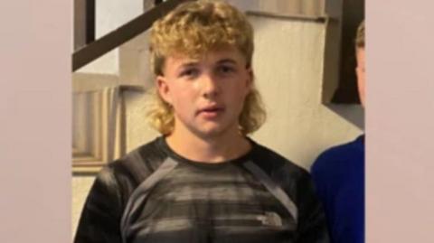A teenager with a blond wavy mullet and a black t-shirt. He is staring directly at the camera.