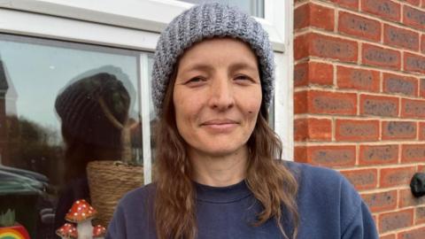 Lucy in a grey hat. She has brown hair and is also wearing a blue jumper. She is stood in front of a window.