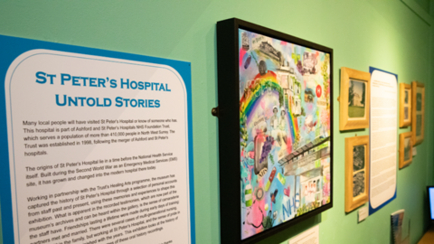 An exhibition on a green wall. On the far left is a poster which reads 'St peter's hospital untold stories' in blue lettering and it has lots of text on it. Next to it is a multi coloured painting, it has graphic including 'NHS' and a rainbow. 