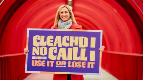 Stiùiriche Seachdain na Gàidhlig, Joy Dunlop, ann an cota dearg agus a' cumail soidhne air a bheil sgrìobhte Cleachd i no Caill i, Use it or Lose it 