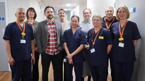 Andrew Henson (third from the left) has brown hair and a brown beard. He has a red and white chequered shirt, black trousers and a smart grey jacket. He is stood with three woman and a man in blue medical uniform and three woman and a man not in uniform. They are standing in a hospital corridor.