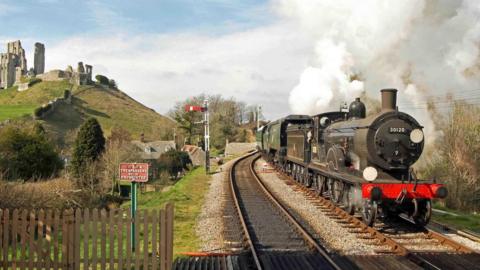 A black steam locomotive travelling past the ruins of Corfe Castle