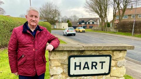 Rob Darby is posing by a stone column with a sign on it that reads: "Hart". He is wearing a red padded coat and has short white hair. He is smiling slightly at the camera.