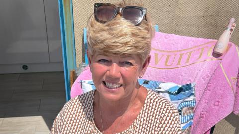 Debbie Booker is sitting outdoors. She has short blonde hair and is wearing sunglasses on top of her head and a patterned shirt. Behind her, a pink towel with the word "VUITTON" is draped over a chair. A bottle is placed on the chair's armrest. The background shows part of an open door and tiled flooring.