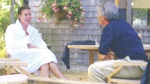 A copy of an undated picture of Peter Mandelson sitting in a white robe laughing while sitting opposite Jeffrey Epstein, who wears a blue top and cream chinos, on a wooden deck.