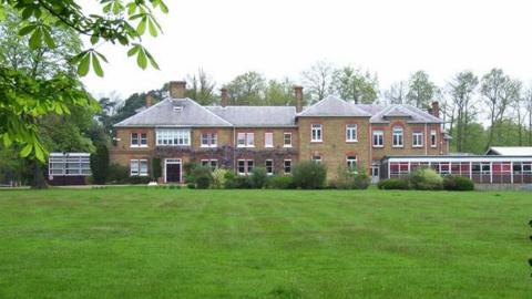 A large school building, with a large green field in front. Next to the main building is a more modern single storey building.