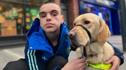 A teenager with short dark hair and brown eyes kneels in the street with an arm around a golden Labrador guide dog. The young man wears a blue Leeds United jacket and the dog has a high visibility harness.