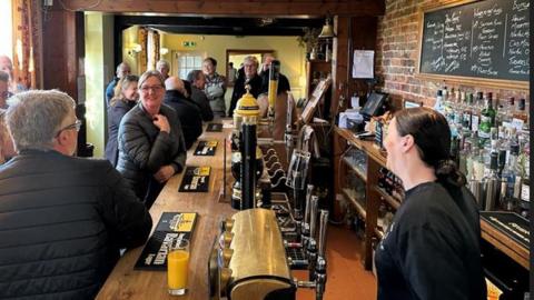 An interior shot of a busy bar area of the Swan on the Green pub in West Peckham with a smiling woman behind the bar serving about half a dozen people sat and propped up the other side of the bar