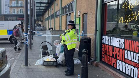 A police officer stands nearby as forensic teams work on a crime scene outside a fast food shop.