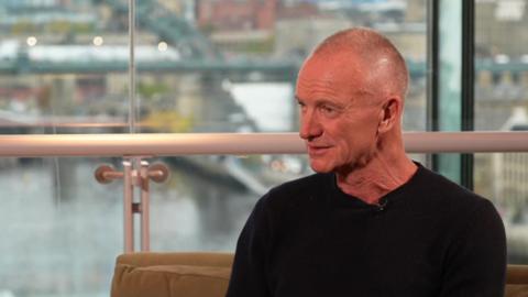 A screenshot of Sting being interviewed. He is sitting on a beige sofa in a building looking out on to the Tyne Bridge. He has a shaved head and he is looking to his right towards the interviewer, who is out of shot. He is wearing a black jumper.