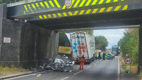 A road under a railway bridge. There is debris under the bridge and two emergency service workers. To the left there is a lorry with its roof damaged.