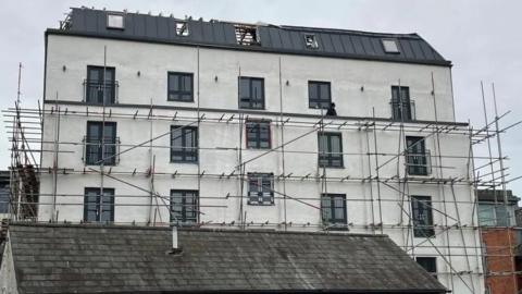 A six storey building with white or cream rendering and scaffolding around it. It is cloudy above.