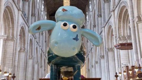A large sculpture of Shaun the Sheep in pale blue with butterflies on its face and ears stands inside Peterborough Cathedral. There are rows of large archways behind the statue. 