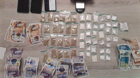 Piles of cash, bags of drugs and several mobile phones are placed on a table.