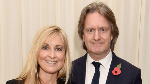 Fiona Phillips and Martin Frizell stand against a beige background. The photo is landscape - both smile and look towards the camera.