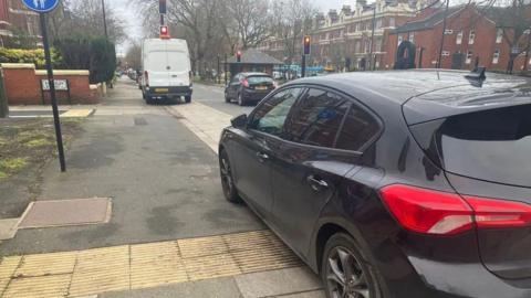 A black car and a white van parked on a pavement in Liverpool near to traffic lights.