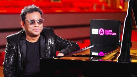 Composer A. R. Rahman sits at a piano, he is wearing a black sparkly jacket and and sunglasses.