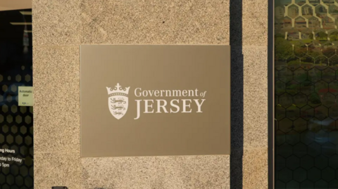 A close-up of the Government of Jersey sign attached to a concrete pillar. On the sign, white text is printed on a brown background.