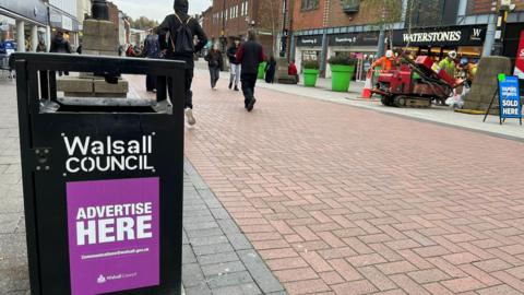 A bin on a pedestrianised street in Walsall. People can be seen walking towards and away from the camera and Waterstones and Superdrug stores are on the right.