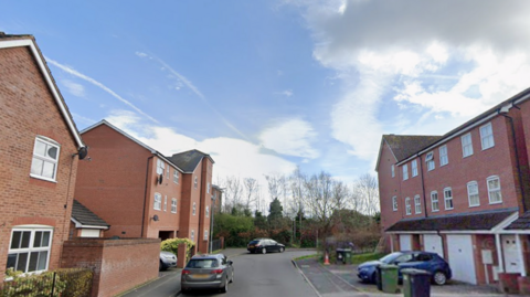 A road with brick houses, parked cars and a cloudy sky.