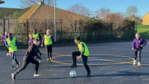 Eight boys and girls playing on a sports pitch, one boy is about to kick a football, four  are wearing high-viz jackets. Four have purple tops on and the rest dark clothing. There are painted circles on the ground, and trees and bushes to the left. 