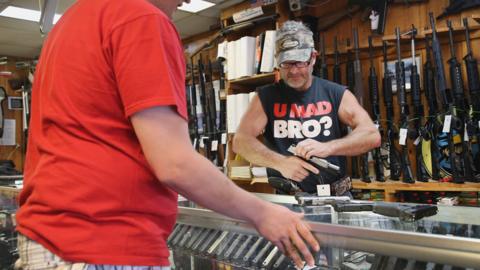 A man tries a gun in a gun store with a t-shirt saying "U mad bro?" and a khaki hat.