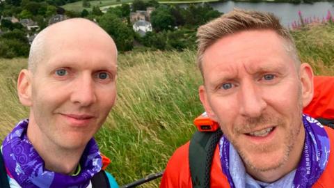 Two men smile for a picture with a green landscape and lake stretching out behind them. The man on the left is shaven, wearing a purple snood. The man on the right also wears a purple snood or scarf, wearing orange clothing.
