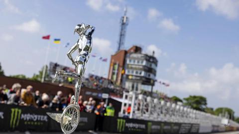 A silver TT trophy in front of the Grandstand with spectators looking on.