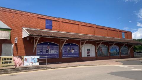 The site of a former Co-op store on Newerne Street in Lydney. The road is in the foreground and in the background, you can see the building, with advertising covering the windows, saying "we care about our customers". Other adverts are visible, including one for ear wax removal and another saying "Lydney pharmacy, open as usual". There are signs of graffiti tagging on one of the hoardings.