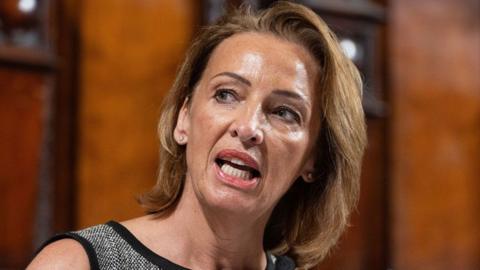 Reform MP Sarah Pochin wearing a sleeveless grey textured top, standing indoors against a dark blurred background with soft lighting