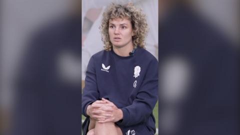 Ellie Kildunne wears a navy blue jumper and short set. She is sitting down and looks past the camera.