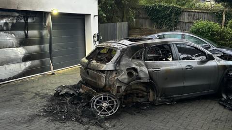 A burnt-out car with large pile of black debris on the ground with a detached wheel. There is a large garage door showing white heat marks and black scorching, and adjacent steps which are also scorched. There is some scorching on the block-paved drive.