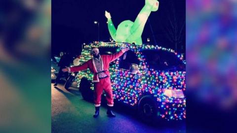 A  man dressed as Santa smiling and posing in front of a van decorated all over with rainbow fairy lights. On top of the car is a lit-up inflatable dinosaur.