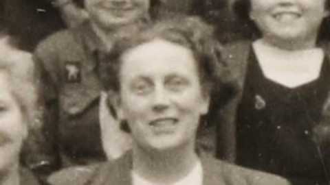 A black and white school photo a a teacher in a blouse and jacket surrounded by children and other staff members.