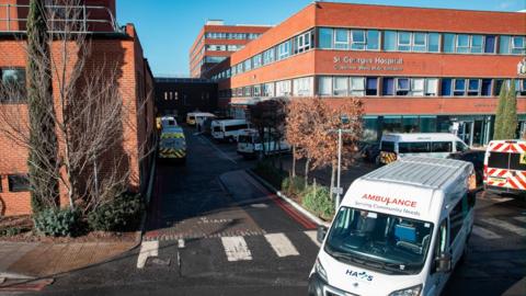 Several ambulances are parked outside St George’s Hospital in Tooting, South London. The large red-brick building has multiple windows and a sign reading ‘Grosvenor Wing Main Entrance’ above the entrance.