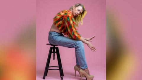 Tom Marshman wearing a multi colour feather half-jumper with denim jeans and cream stiletto heels. He is sat on a brown stool with his hands out in front, looking at the camera with a pink studio backdrop.