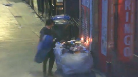 A CCTV image shows Daniel Byrne standing next to a large sack of rubbish in front of the doors of the Beatles Museum. The rubbish is starting to burn, and flames can be seen rising from it. 