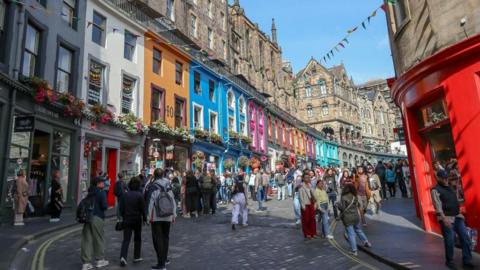 A busy cobbled street with colourful shop fronts in the background and hundreds of people milling about