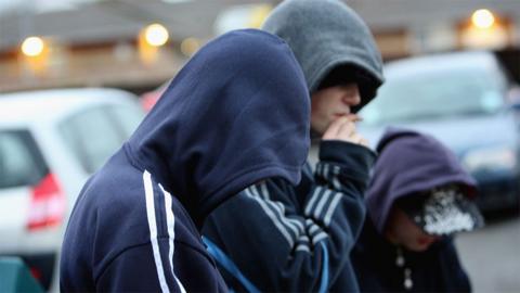 Generic close up image of three young people in hooded tops so their faces can't be seen. They are facing to the right and the middle one is smoking