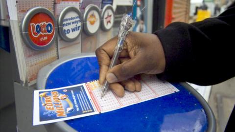 Photo of a hand filling out a EuroMillions ticket at a booth in a corner shop