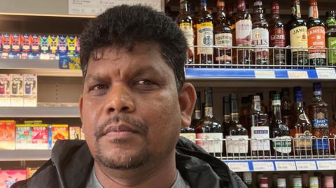 Man looking into the camera in a shop, with colourful bottles on display behind him. He has swept-back black hair and a neat moustache and beard.