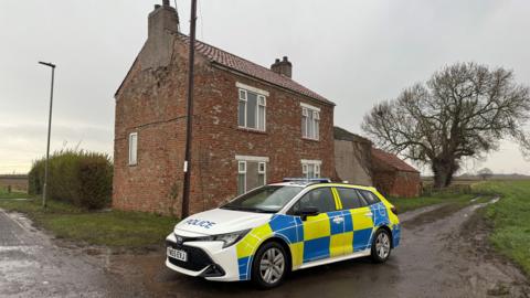 A marked police car parked on a country lane in front of a redbrick house in the countryside. The sky is overcast.